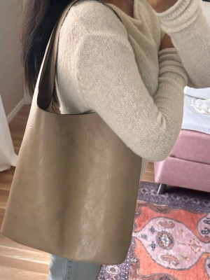 Beige axelväska i konstläder - Stilren beige axelväska i mjukt konstläder med enkel bucket-form och bred axelrem. Väskan är rymlig och har en minimalistisk design utan synliga detaljer eller loggor. Perfekt för dig som gillar en clean och modern look.