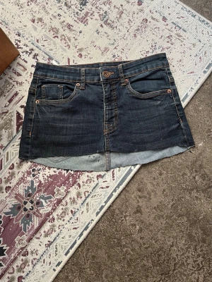 Mörkblå jeanskjol med fickor - Säljer en kort mörkblå jeanskjol med klassiska fem fickor och knappstängning fram. Kjolen har rå kant nedtill för en avslappnad look och är gjord i denim. Perfekt till sneakers eller boots. Klippt