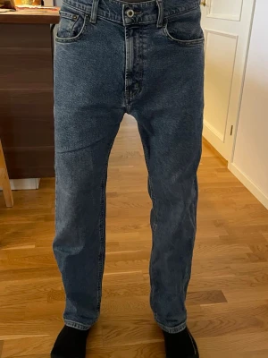 Blå raka jeans med hög midja - Säljer ett par klassiska blå jeans med rak passform och hög midja. Jeansen har fem fickor, bälteshällor och stängs med knapp och dragkedja. Materialet är kraftig denim i bomull och färgen är en tvättad blå nyans.