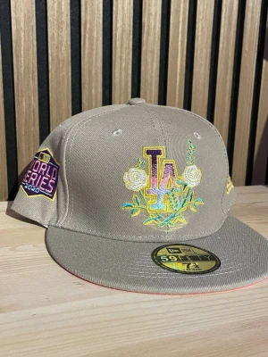 Beige LA World Series 2020 keps - Snygg beige keps med broderad LA-logga i gult, lila och vitt framtill samt blommotiv. På sidan finns ett färgglatt World Series 2020-märke. Platt skärm och klassisk 59FIFTY-modell. Perfekt för dig som gillar sportig streetstyle.