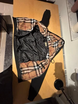Burberry hoodie  - Svart hoodie med dragkedja och huva. Insidan av huvan och jackan har ett klassiskt beige, svart och röd rutigt mönster. Utsidan är helsvart med ribbade muddar och fickor framtill. Perfekt för en stilren och trendig look.