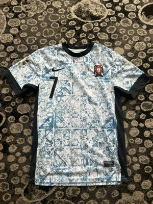 Portugal bortatröja Nike nr 7 - Portugal bortatröja från Nike med nummer 7. Tröjan är vit med blått abstrakt mönster, svarta detaljer och rund hals. Portugal-emblemet på bröstet och Nike-logga. Korta ärmar och lätt, andningsbart material perfekt för fotboll.