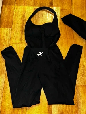 Svart halterneck jumpsuit - Snygg svart jumpsuit med halterneck och figurnära passform. Jumpsuiten har en markerad byst och ett litet vitt X-tryck framtill i midjan. Perfekt för dig som gillar en stilren och trendig look. Materialet ser stretchigt ut för extra komfort.