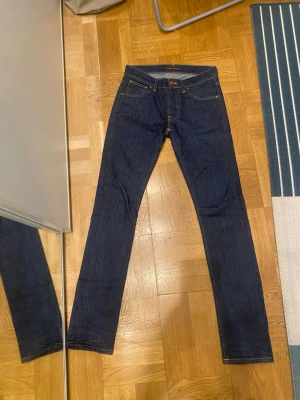 Mörkblå jeans från Nudie Jeans - Säljer ett par mörkblå jeans från Nudie Jeans med klassisk femficksmodell och raka ben. Jeansen har kontrastsömmar i orange och är tillverkade i kraftig denim. Perfekta för dig som gillar stilrena och tidlösa jeans.(Står L 34 men passar L 32).storlek W28 L 32