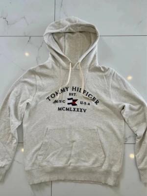 Ljusbeige hoodie från Tommy Hilfiger - Ljusbeige hoodie med broderad Tommy Hilfiger-logga och flagga på bröstet. Perfekt för en chill och stilren look. Materialet är mjukt och bekvämt, passar till jeans eller joggers.