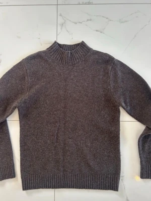 Brun stickad tröja med hög krage - Brun grå stickad tröja från Sandö med hög ribbad krage och långa ärmar. Tröjan har ribbade muddar vid ärmslut och nederkant. Perfekt nu för vintern och kyligare dagar. Aldrig använts men har inte prislappen kvar