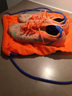 Nike Mercurial Elite vapor 14 - Säljer ett par Exclusiva Nike Mercurial fotbollsskor i orange med gröna detaljer och snörning. Skorna har en sportig, strömlinjeformad design och är tillverkade i ett lätt syntetmaterial för bästa känsla på planen. Perfekta för dig som vill sticka ut på fotbollsplanen.