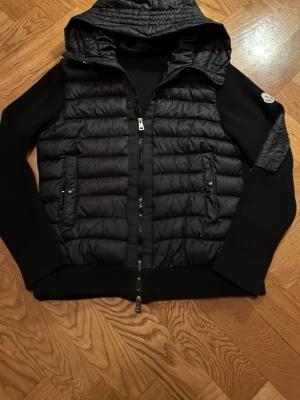 Svart cardigen med luva - Snygg svart cardigen med quiltad front och luva. Jackan har dragkedja framtill, två fickor med knappar och släta ärmar i mjukt material. Perfekt för kyliga dagar och har en modern, sportig look.