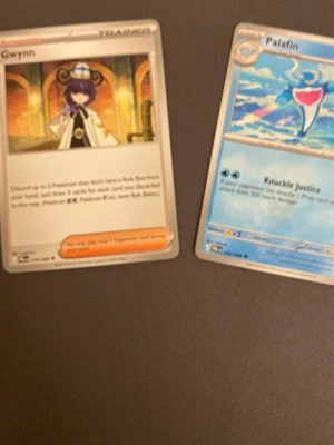 Pokémonkort Palafin & Gwynn - Två Pokémonkort: Palafin med blå och vita färger och 150 HP, samt tränarkortet Gwynn med en karaktär i vit mantel. Båda korten har klassisk rektangulär form och är tryckta på glansigt papper. Perfekt för samlare eller Pokémon TCG-spelare.