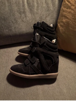 Isabel marnat  - Säljer ett par svarta isabel marant med kilklack och kardborreband. Skorna har en cool design med paneler i mocka och textil, samt vit sula som kontrast. Perfekta för dig som vill ha både stil och höjd i ett par sneakers. Har även kvitto som jag kan skicka bild på kommer även med dustbag