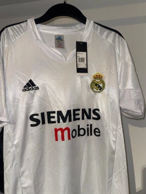 Real Madrid Zidane matchtröja Adidas - Klassisk vit Real Madrid matchtröja från Adidas med svarta detaljer och klubbmärke på bröstet. Siemens Mobile som sponsortryck och Zidane med nummer 5 på ryggen. Tillverkad i glansigt funktionsmaterial, perfekt för fotboll eller samling.