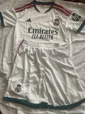 Real Madrid matchställ Adidas - Säljer ett Real Madrid matchställ från Adidas i vitt med gröna och vinröda detaljer. Tröjan har klubbmärke, Adidas-logga och Emirates Fly Better-tryck. Shortsen har klubbmärke och gröna detaljer. Perfekt för fotbollsträning eller match!