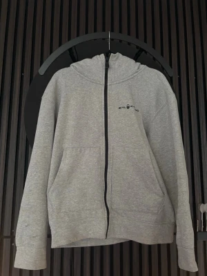 Grå zip hoodie från Sail Racing - Grå hoodie från Sail Racing med dragkedja framtill och svart logga på bröstet. 