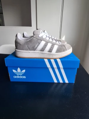 Adidas grå sneakers i mocka - Gråa Adidas sneakers i mocka med klassiska vita ränder på sidorna och vit sula. Skorna har lågt skaft och vit snörning. Yttersulan är i gummi med fiskbensmönster. Perfekta för en stilren och sportig look. Bara några fläckar på mackan men det ör det som gör dom så snygga.