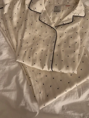 Vit prickig pyjamas med svarta prickar - Säljer en vit pyjamas med små svarta prickar och svart detaljer. Setet består av en skjorta med krage och knappar samt byxor med resår i midjan. Materialet är mjukt och lent. 