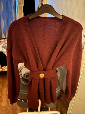 Vinröd kofta - Snygg vinröd omlottkofta med bred ribbad kant och matchande bälte med dekorativ knapp i midjan. Koftan har lång ärm och en mjuk, stickad känsla som ger en avslappnad men trendig look. 