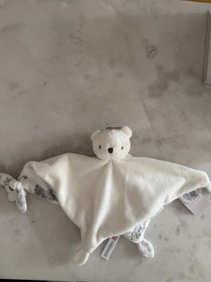 Vit snuttefilt med björnhuvud - Mysig snuttefilt i mjuk vit plysch med ett gulligt björnhuvud och knutar i hörnen. Filten har detaljer i grått med djurmönster på insidan. Perfekt som trygghet för små barn.