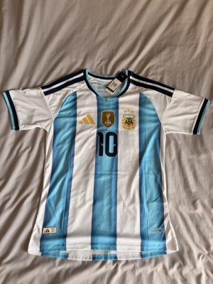 Argentina Messi matchtröja blåvit - Säljer en officiell Argentina matchtröja från Adidas med Messi och nummer 10 på ryggen. Tröjan har klassiska blå och vita ränder, svarta detaljer och guldiga loggor på bröstet. Tillverkad i lätt och ventilerande material, perfekt för fotboll eller träning.
