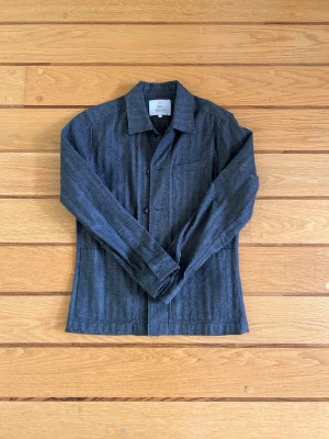 Mörkgrå overshirt från Bläck - Stilren mörkgrå overshirt från Bläck i fiskbensmönstrad bomull. Skjortjackan har klassisk krage, knappstängning framtill och en bröstficka. Perfekt lager på lager-plagg för en clean och modern look.