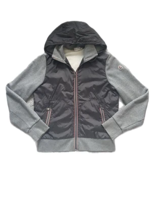 Moncler Cardigan Grå - Snygg Moncler Cardigan i grått och svart med huva och dragkedja framtill. Cardiganen har gråa ärmar i sweatshirtmaterial och svart glansig kropp. Två fickor med dragkedja och ribbade muddar. Perfekt för lager på lager under kyliga dagar. Storlek M men passar S. Köpt från Von Greven Closet