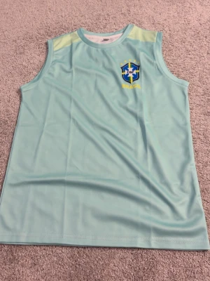 Ljusblå Brasilien ärmlös träningströja - Snygg ljusblå ärmlös träningströja med Brasilien-logga och texten 'Brasil' på bröstet. Tillverkad i ett lätt och snabbtorkande material, perfekt för träning eller sportaktiviteter. Tröjan har rund hals och ljusgula detaljer vid axlarna.