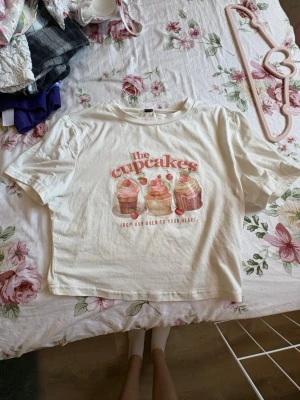 Vit t-shirt med cupcake-tryck - Söt vit t-shirt med ett färgglatt cupcake-tryck och texten 'the cupcakes' framtill. T-shirten har korta ärmar och en avslappnad passform. Perfekt för dig som gillar gulliga motiv och vill ha en unik look.
