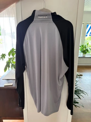 Grå och svart träningströja med dragkedja - Snygg långärmad träningströja i grått och svart med halv dragkedja framtill. Tillverkad i ett lätt och stretchigt funktionsmaterial som andas, perfekt för löpning eller gym. Raglanärmar för extra rörelsefrihet och sportig look.
