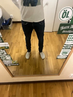 Mörkblå nudie slim fit jeans - Säljer ett par mörkblå slim fit jeans från nudie. Jeansen har en snygg passform och detaljerade bakfickor med broderad våg. Perfekta för dig som gillar en stilren och enkel look. Jeansen har även snusfade på backfickan jag är 178 och väger ich 60 kg storlek 32:32 siter tajt på mig som du ser på bilderna