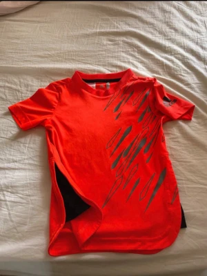 Röd Adidas t-shirt med tryck - Cool röd/orange  t-shirt från Adidas med svart grafiskt tryck på framsidan. T-shirten har korta ärmar, rund halsringning och en unik slitsdetalj på sidan