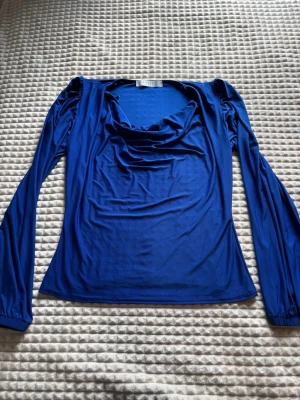 Blå långärmad topp med drapering - Snygg blå långärmad topp med draperad urringning och puffade axlar. Tillverkad i ett mjukt och stretchigt material som ger en smickrande passform. Perfekt att styla med jeans eller kjol för en trendig look.
