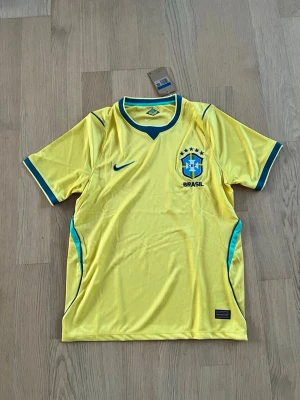 Gul Brasil Nike fotbollströja - Säljer en gul Brasilien fotbollströja från Nike med blå detaljer och gröna inslag. Tröjan har korta ärmar, rund halsringning och Brasil-logga på bröstet. Perfekt för dig som älskar fotboll och vill stötta landslaget.