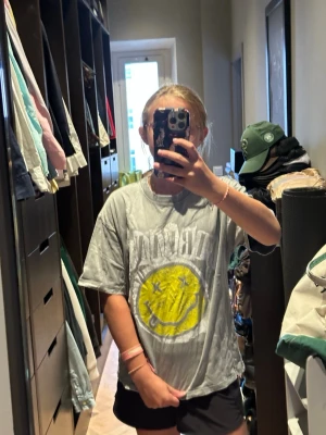 Grå Nirvana t-shirt med tryck - Säljer en grå t-shirt med stort Nirvana-tryck framtill och det klassiska gula smiley-ansiktet. T-shirten har korta ärmar och rund hals. Perfekt för dig som gillar bandtröjor och vill ha en avslappnad stil.