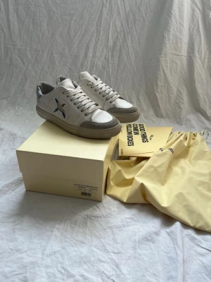 Vita axel arigato skor  - Snygga vita sneakers med gråa och beige inslag samt en blå detalj på sidan. Skorna har klassisk låg profil, rund tå och vita snören. Materialet ser ut att vara en mix av läder och mocka. Perfekta för en clean och stilren look.