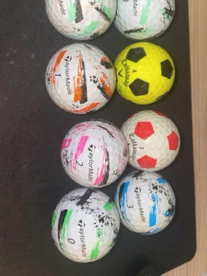 Golfbollar TaylorMade & Callaway mix - Säljer ett set med åtta golfbollar från TaylorMade och Callaway. Bollarna har olika färgmarkeringar i grönt, orange, rosa, blått och svart samt två bollar med fotbollsmönster i gult och vitt med svarta och röda detaljer. Perfekt för dig som vill ha unika bollar på golfbanan.