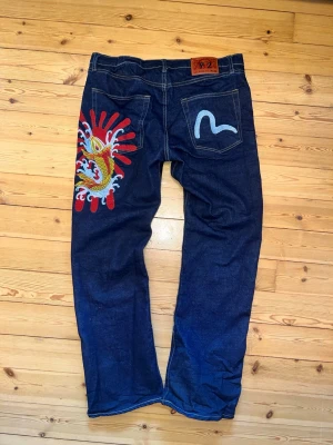 Mörkblå jeans med drakbrodyr - Unika mörkblå jeans i rak modell med färgglad drakbrodyr på ena bakfickan och kontrastsöm. Jeansen har klassisk femficksdesign och en patch i läderimitation i midjan. Materialet är kraftig denim i bomull. Perfekta för dig som vill sticka ut med coola detaljer.