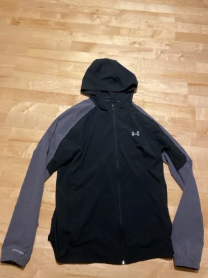 Under Armour svart/grå vindjacka - Sportig vindjacka från Under Armour i svart med grå ärmar och huva. Jackan har hel dragkedja, logga på bröstet och texten 'HeatGear' på ärmen. Tillverkad i lätt syntetmaterial som andas, perfekt för träning och utomhusaktiviteter.