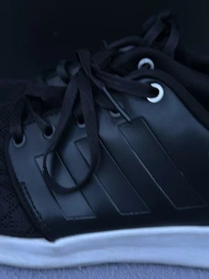Svarta Adidas sneakers med vita sulor - Säljer ett par svarta Adidas sneakers med klassiska tre ränder på sidan och vita sulor. Skorna har svarta skosnören och detaljer i vitt på hälen där Adidas-loggan syns tydligt. Materialet är en mix av syntet och textil.   Som nya knappt använda    Stl 37,5