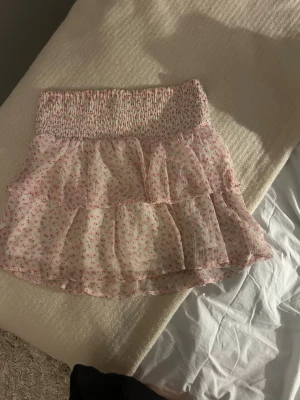 Kort volangkjol med blommigt mönster - Söt kort kjol med volanger och smock i midjan. Kjolen är ljusrosa med små blommor i rosa och grönt. Tillverkad i ett lätt, luftigt material som passar perfekt till varmare dagar.