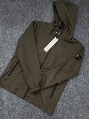 Stone Island Windbreaker  - Snygg olivgrön vindjacka från Stone Island med huva och svarta dragkedjor. Jackan har två sidofickor med dragkedja, elastiska muddar vid ärmsluten och en patch på vänster ärm. Perfekt för blåsiga dagar och enkel att matcha med streetwear.