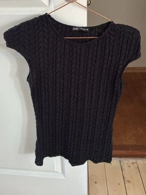 Svart ribbad topp från Zara - Säljer en svart ribbad topp från Zara med korta ärmar och rund hals. Toppen har ett snyggt stickat mönster som ger en cool struktur och sitter tight mot kroppen. Perfekt att styla med jeans eller kjol för en trendig look.