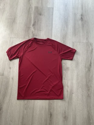 Röd tränings-tshirt från Under Armour - Snygg röd tränings-tshirt från Under Armour med korta ärmar och rund hals. Tillverkad i ett lätt och snabbtorkande funktionsmaterial som passar perfekt till gymmet eller löprundan. Diskret logga på bröstet.