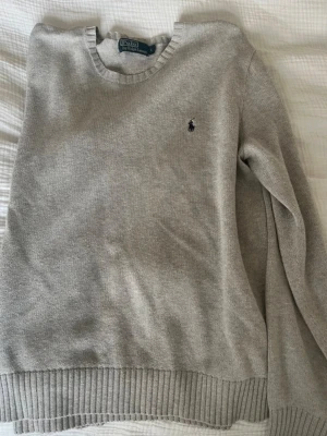 Ljusgrå stickad tröja från Polo Ralph Lauren - Ljusgrå stickad tröja från Polo Ralph Lauren med rund halsringning och klassisk marinblå logga broderad på bröstet. Tröjan har ribbade muddar vid ärmslut och nederkant. Perfekt för lager på lager och enkel att matcha.