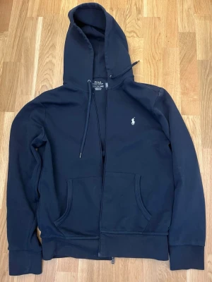 Mörkblå full zip från Polo Ralph Lauren - Klassisk mörkblå tröja från Polo Ralph Lauren med dragkedja, fickor framtill och justerbar huva. Broderad logga på bröstet och ribbade muddar vid ärmslut och nederkant. Tillverkad i mjuk bomullsblandning för extra komfort.