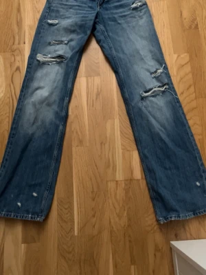 Blå slitna raka jeans - Säljer ett par blå jeans med raka ben och flera slitningar och hål på både fram- och baksida. Jeansen har en avslappnad passform och är tillverkade i klassiskt denimtyg. Perfekta för en trendig och avslappnad stil.