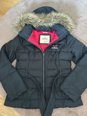 Mörkblå pufferjacka från Hollister  - Otroligt snygg mörkblå pufferjacka från Hollister med fuskpäls på huvan och rosa innerfoder. Jackan har bälte i midjan, dragkedja framtill och broderad logga på bröstet. Perfekt för kalla höst/vinterdagar. Jackan är från 2012 så finns inte att köpa idag så den är väldigt rare. Storlek M.