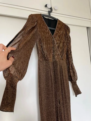 Glittrig brun långklänning med v-ringning - Säljer en glittrig guld/brun jumpsuit med v-ringning och långa puffärmar. Klänningen har omlottdetalj framtill och är gjord i ett skimrande, lätt material som ger en lyxig och glittrig känsla. Perfekt för dig som vill sticka ut med metallic vibes och coola detaljer. Materialet är stretchiga och passar xs, s eller M 