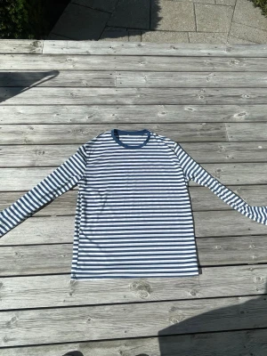 Randig långärmad tröja blå/vit - Säljer en långärmad tröja med klassiska marinblå och vita horisontella ränder. Tröjan har rund halsringning med blå kant och är gjord i ett mjukt, tunt material som passar perfekt till jeans eller kjol.