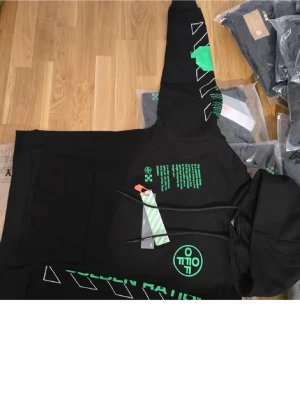 Svart hoodie med grönt tryck - Svart hoodie med stora gröna och vita tryckdetaljer på både fram- och baksida. Har huva med snören och en stor magficka. Trycken består av text, symboler och grafiska element som ger en modern streetwear-känsla.