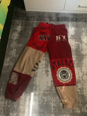 Patchwork mjukisbyxor med NYC-tryck - Unika mjukisbyxor i patchwork-stil med röda och beige partier samt stora tryck med NYC, ATHLETIC och college-tema. Byxorna har resår i midja och benslut, samt flera coola detaljer och texttryck som ger en sportig och trendig vibe.