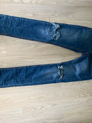 Blå slitna jeans med hål - Säljer ett par blå jeans med skinny fit och slitna detaljer på båda benen. Jeansen har stora hål vid knäna och en tvättad look som ger en cool och avslappnad stil. Perfekta för dig som gillar streetwear och trendiga outfits.
